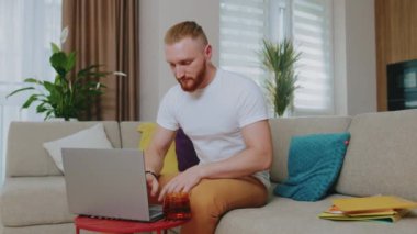 Odaklanmış genç adam kanepede otururken dizüstü bilgisayar kullanıyor. Çay iç. Evde çalış. İnternet teknolojisi. Karantina zamanı. Kapat..