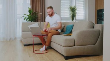 Evde oturan yakışıklı genç adam kanepede otururken dizüstü bilgisayar kullanıyor. Çay iç. Çalış. İnternet teknolojisi. Karantina zamanı. Kapat..