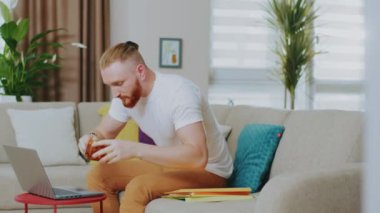 Kanepede oturan sinirli, genç ve yakışıklı bir adam dizüstü bilgisayar kullanıyor. Çay iç. Evde çalış. İnternet teknolojisi. Karantina zamanı. Kapat..