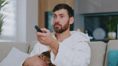 Kedili genç adam televizyon izliyor. Kanepede oturup gülümsemek. Dinlenme sinema kanalları dinleniyor. Portre. Kapatın. Yavaş çekim.