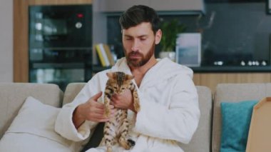 Yakışıklı genç adam kediyle oynuyor. Kanepede oturup gülümsemek. Dinlenme sinema kanalları dinleniyor. Portre. Kapatın. Yavaş çekim.