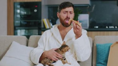 Kedisi olan genç duyarlı bir adam televizyon izlerken telefon kullanıyor. Kanepede oturup evde hüzünle ağlamak. Dinlenme sinema kanalları dinleniyor. Portre. Kapatın. Yavaş çekim.