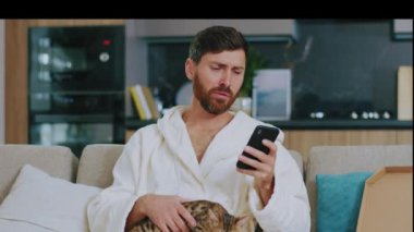 Ağlayan, kedisi olan hassas bir adam televizyon izlerken telefon kullanıyor. Evdeki kanepede üzgün oturmak. Dinlenme sinema kanalları dinleniyor. Portre. Kapatın. Yavaş çekim.