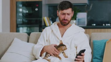 Kapat çeneni, kedisi olan hassas adam TV izlerken telefonu kullanıyor. Evdeki kanepede üzgün oturmak. Dinlenme sinema kanalları dinleniyor. Portre. Yavaş çekim.