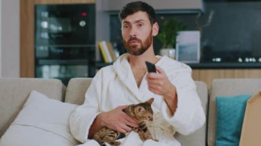 Ciddi, çekici ve kedisi televizyon izleyen genç bir adam. Evdeki kanepede oturmak. Dinlenme sinema kanalları dinleniyor. Portre. Kapatın. Yavaş çekim.