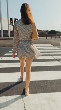 Güzel çiçekli bir elbise giymiş şık bir kadın, parlak güneş ışığı altında işlek bir yaya geçidinde güvenle yürür.