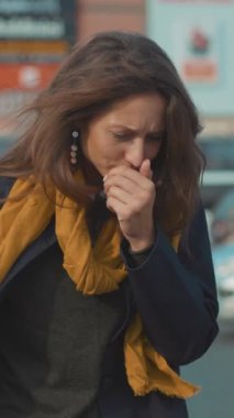 Düşünceli bir kadın şehir hayatının ortasında duruyor, derin düşüncelerinde kaybolmuş, rahat bir eşarba sarılmış.