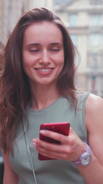 Neşeli genç bir kadın, şehir ortamının canlı atmosferinin tadını çıkarırken akıllı telefonuyla mutlu mesut meşgul oluyor.