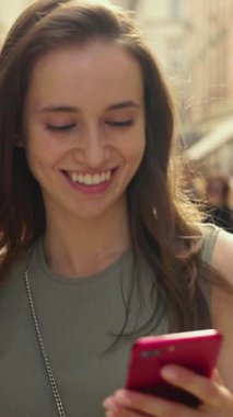 Neşeli genç bir kadın canlı ve canlı bir şehirde akıllı telefonuna bakarken parlak bir gülümseme sergiliyor.