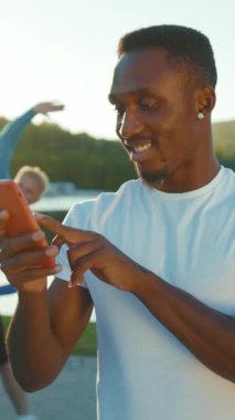 Gülümseyen, enerjik genç bir adam açık havada egzersiz yaparken akıllı telefonuyla neşeyle meşgul olur.