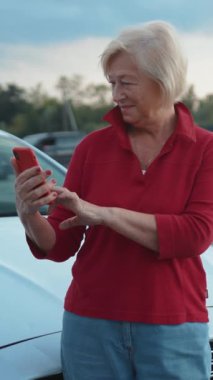 Akıllı telefonuyla meşgul yaşlı bir kadın güpegündüz park halindeki bir arabanın yanında sevinçle gülümsüyor.