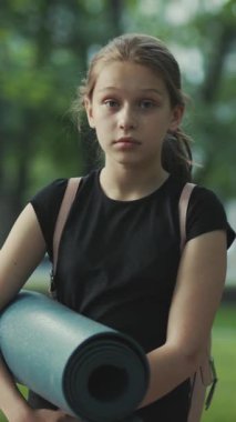 Siyah kıyafetli düşünceli genç bir kız. Yoga minderi taşıyor. Etrafı yemyeşil.