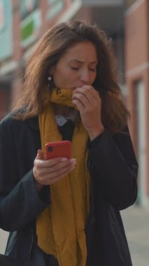 Düşünceli genç bir kadın, hareketli bir şehir ortamında yürürken akıllı telefonunu dikkatle kontrol ediyor.