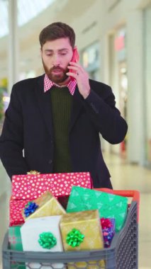 Şık bir adam alışveriş merkezinde alışveriş yapıyor. Etrafı canlı, renkli hediyeler ve dekorasyonlarla çevrili.