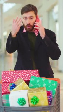 Bir adam renkli tatil hediyeleri ve süslemelerle dolu bir arabayı iterken telefonda konuşuyor.