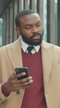 İyi giyimli bir iş adamı modern bir şehir içinde yürürken akıllı telefonunu kontrol ediyor.
