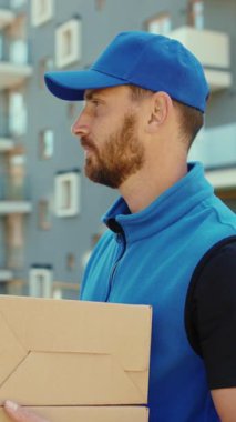 Mavi şapkalı ve ceketli neşeli bir teslimatçı karton bir kutu getirirken gülümsüyor, dostane tavrını sergiliyor.