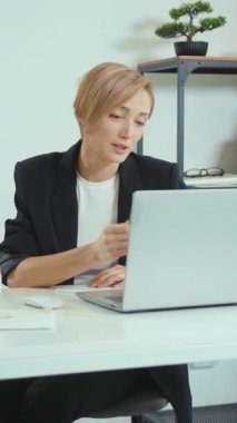 Sıcak bir ofiste, bir kadın dizüstü bilgisayarı ve notları arasında birden fazla iş yapar, modern bir çalışma alanında üretkenlik ve yaratıcılık sergiler.