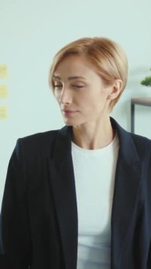 Siyah ceketli ve beyaz gömlekli kendine güvenen bir kadın modern bir ofiste kameraya sarılıp profesyonellik ve pozitiflik sergiliyor.