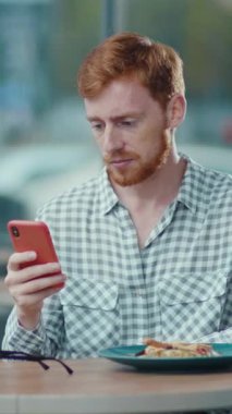 Bir adam masada oturuyor ve dikkatle cep telefonuna bakıyor, ekranda gösterilen şeye odaklanmış görünüyor.