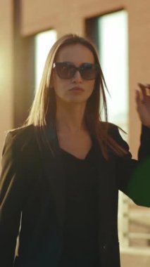 Şık siyah ceketli ve güneş gözlüklü kendine güvenen bir kadın cep telefonuyla bir şehir binasının önünde konuşuyor.