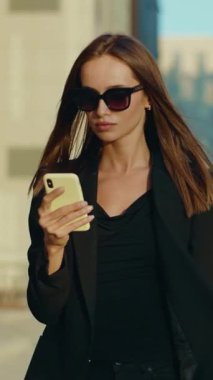 Şık siyah ceketli ve şık güneş gözlüklü bir kadın, şehir ortamında cep telefonuyla iletişim kurarken izleyicileri kendine güveniyle büyülüyor.