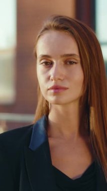 Uzun kahverengi saçlı, kendine güvenen bir kadın şık bir siyah ceket ve bluzla ortaya çıkar. Kamerayla çekim yaparak güç ve zarafet sağlar.