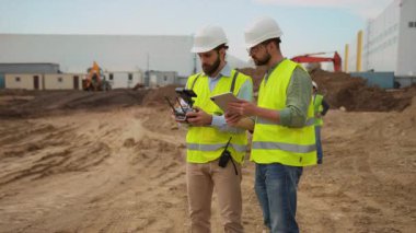 Uçan İHA 'yı kontrol ederken ellerinde tablet ve akıllı telefon tutan parlak sarı yelekli iki müteahhit. Topograflar arka planda. Açık havada.