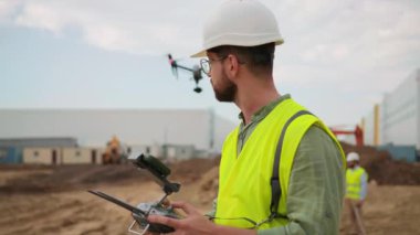 Sarı yelek ve beyaz başlıklı genç erkek işçinin inşaat alanında kuadkopter kullanırken akıllı telefon ekranına bakışının yan görüntüleri. Foreman uzaktan kumandalı pilotsuz uçak kullanıyor.