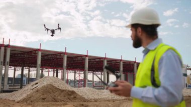 Sarı yelek ve beyaz şapka giyen ciddi genç erkek işçinin gökyüzünde kırmızı ışık saçan uçan bir drona bakışını çekmek. Quadcopter kullanan bir adamın görüntüsü. İnşaat işi