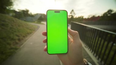 Kırsal bölgedeki köprünün yanında yürürken telefon kullanan tanınmayan genç bir adam. Yeşil ekranlı, erkek el ele tutuşan telefonun yakın çekimi. Akıllı telefondan mesaj okuyan adam. Akşam vakti. Dışarıda.