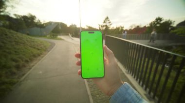 Akşamleyin köprünün yanında yeşil ekrana dokunan, akıllı telefonu tutan bir erkeğin görüntüsü. Özel bölgede yürürken telefonu kontrol eden adam. Halk parkında dinleniyor. Açık havada.