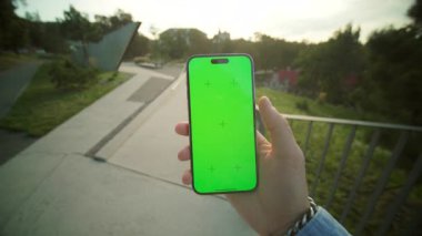 Siyah akıllı telefonları yeşil ekran ile tutan erkeklerin yakın çekim görüntüleri. Genç adam cep telefonu ve maket kullanıyor. Arkaplanda çağdaş parkın bulanık görünümü