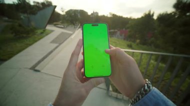 Yeşil ekranı gösteren telefonları olan yakın plan erkek elleri. Genç bir adam akşam parkında yürürken arkadaşıyla mesajlaşıyor. Arka planda eski şehir mimarisi, dışarıda. Yaz zamanı. Krom anahtar kavramı. Dışarıda.
