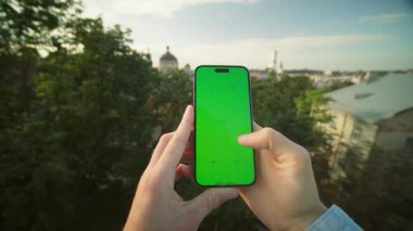 Erkek ellerinin akıllı telefondaki yeşil ekrana dokunuşunu yakından çek. Genç bir adamın cep telefonuyla maket yaptığını gösteren yatay görüntü. Bulanık arka planda eski şehir binaları. Açık havada. Teknoloji ve modern