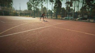 Kendini işine adamış bir oyuncu, verimli ağaçlarla çevrili, güneşli bir tenis kortunda çeşitli çalışmalar yapıyor.