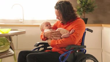 Tekerlekli sandalyedeki genç babanın portresi. Kahvaltıdan sonra mutfakta tatlı bir bebek dinleniyor. Engelli biri. Sevgi, ilgi. Aile kavramı. Babalık. Babalık