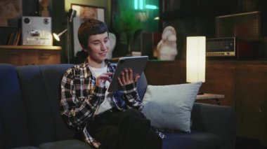 İnternette gezinen kaygısız genç bir kadını çekmek, videodan zevk almak. Kareli gömlekli neşeli, barışçıl bir kız kanepeye oturmuş, elinde dijital tabletle kanepede oturuyordu.