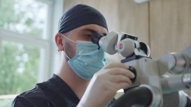 Mavi üniformalı ve maskeli genç bir doktor dişçi ameliyat mikroskobuna yüksek çözünürlüklü bakıyor. Tıp konsepti. Tedavi için. Diş hekimliği. İçeride. Gündüz