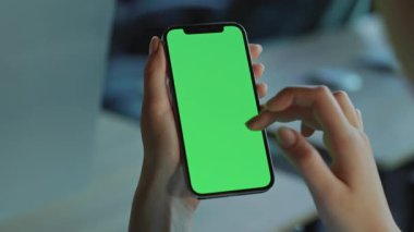 Yeşil ekranlı dikey akıllı telefon kullanan bir kadın. Modern cep telefonunu sol elinde tutan kadın eller, bulanık arka planda internette sörf yapıyor. İçeride. Gadget, teknoloji