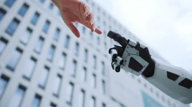 İnsan elinin altından ve robot elin parmak uçlarını göstererek dışarı bak. Arka planda iş merkezinin bulanık görüntüsü. Beyaz modern bina. Gündüz vakti. Şehre. Fütürist. Şehir tasarımı. Yapay