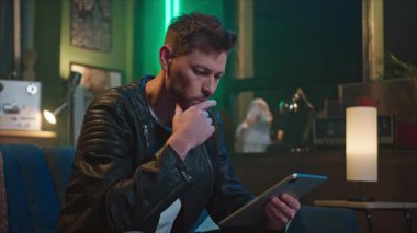 Düşünceli, şık kıyafetli genç bir adamın tablet ve dokunmatik ekran kullanarak koltukta oturduğu görüntüler. Mesaj yazan adam. İçeride. Akşam vakti. Evden çalış.