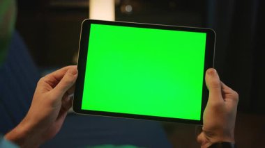 Erkek eller dijital tableti tutuyor ve akşam vakti evin arka planında yeşil ekran var. Modern teknolojiler, cihazlar. Kapalı