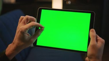 Kanepede oturan ve akşamları evde boş yeşil ekran tablet kullanan tanınmayan bir adamın yakın çekim görüntüleri. Parmaklar ekranı büyütmek için çimdikliyor. Kapalı