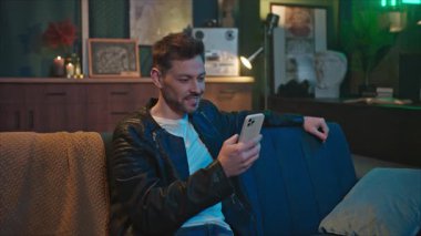 Kanepede oturan ve oturma odasında akıllı telefon kullanan gülümseyen yakışıklı adamın görüntüsü. Genç, rahat, beyaz bir adam akşamları ekrana dokunuyor ve zoom yapıyor. Kapalı