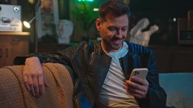 Kanepede oturan, elinde cep telefonu olan ve ekrana bakan mutlu bir genç adam. Akıllı telefondan mesaj atan şık bir adam. İnternet, uygulamalar. Akşam evde dinlen.
