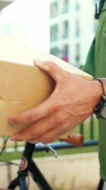 Yeşil kıyafetli bir teslimatçı elinde kahverengi bir kutuyla dışarıda duruyor. Şehir lojistik operasyonlarını temsil ediyor.