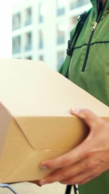 Yeşil ceketli bir teslimatçı gün boyunca kalabalık bir şehir caddesinde karton bir kutu tutuyor.