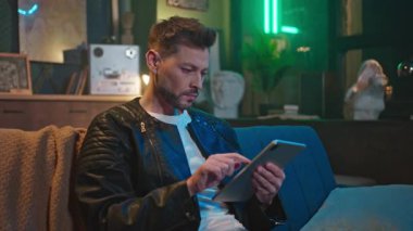 Deri ceketli, düşünceli bir gencin kanepede oturup oturma odasında internette sörf yaptığı görüntüler. Tablet kullanarak iş problemini çözen yorgun adamın portresi. Modern iç mimari