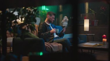 Kanepede oturan, çay içen ve akşamları tablet kullanan yakışıklı, odaklanmış beyaz bir adamın yatay çekimi. Dairenin içi çok güzel. Kapalı